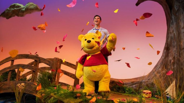 Winnie the Pooh puppet and puppeteer on a stage with falling leaves and some toys.