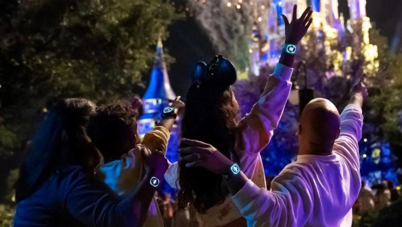 A family holds illuminated MagicBand+ in the air as fireworks explode over Cinderella Castle